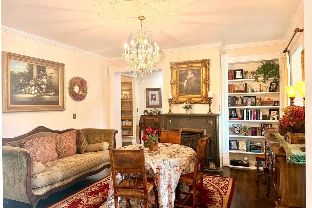Ein Essbereich mit einer Chandelüster, einer Couch, einem Tisch mit Blumenmuster-Tischdecke und einem Bücherregal mit gerahmten Gegenständen.