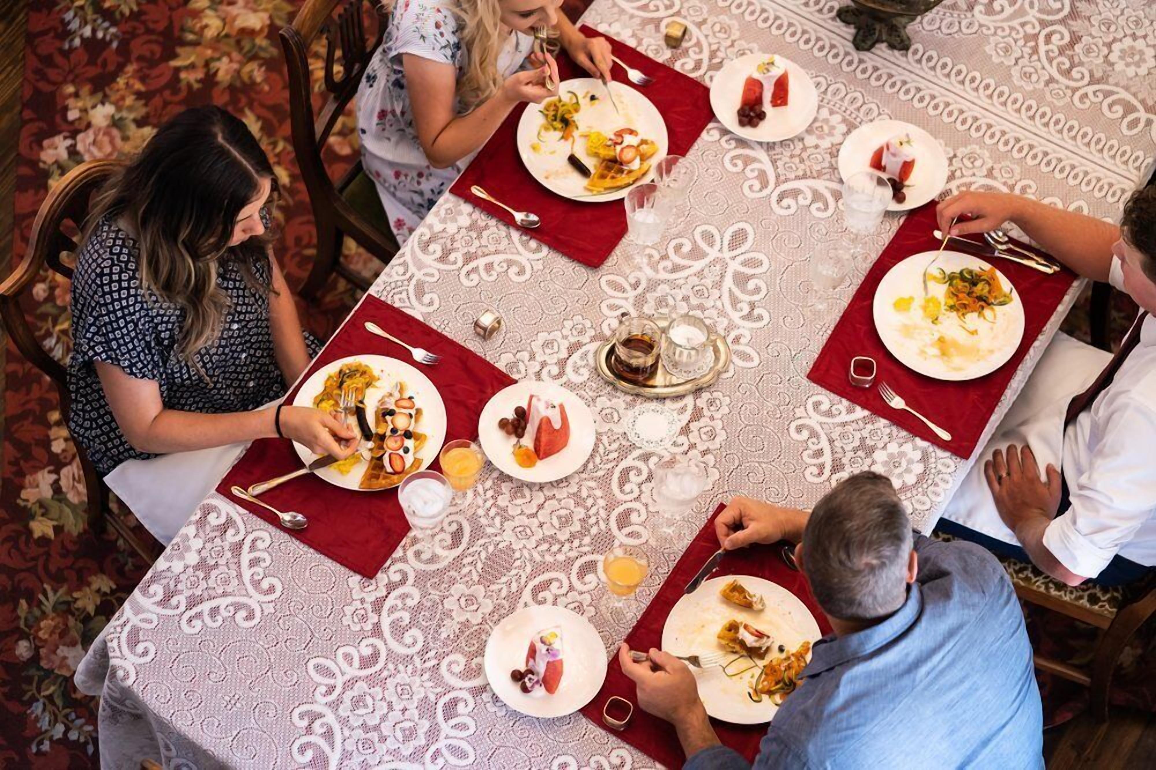 Un groupe de personnes est assis à une table, devant un repas, et la table est recouverte d’une nappe à motifs. Des chaises sont disposées autour de la table.