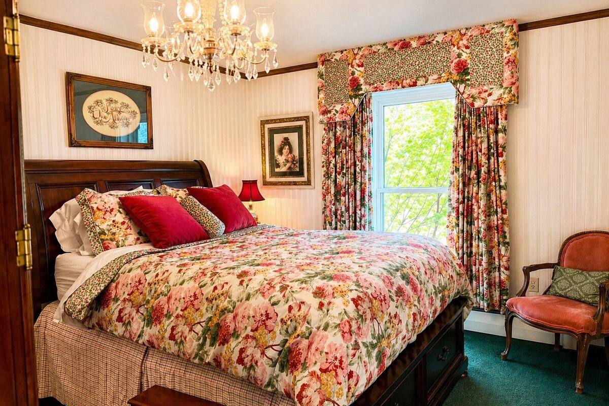 Un dormitorio con colcha estampada, cabecera de madera, araña y ventana con cortinas florales, además de una silla.
