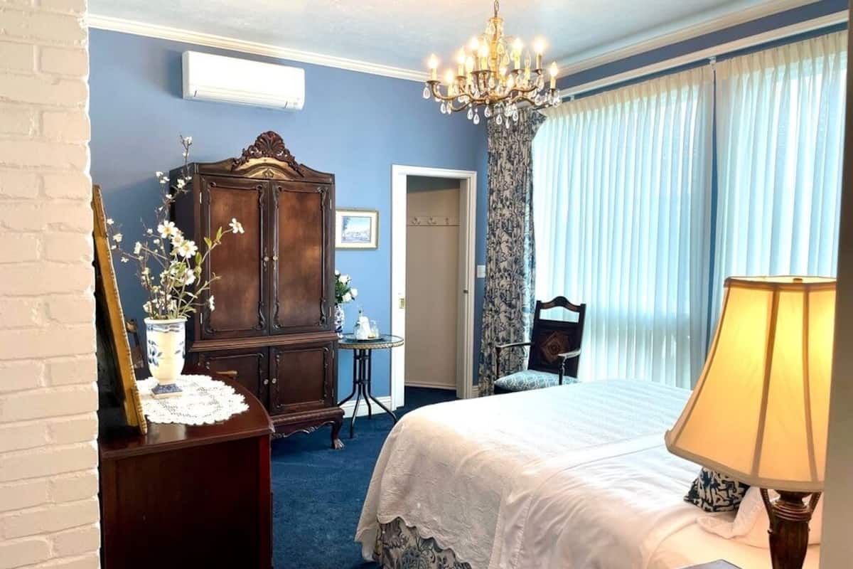 Un dormitorio con cama, candelabro, cómoda, espejo, una mesa con un florero y una puerta que da a otra habitación.