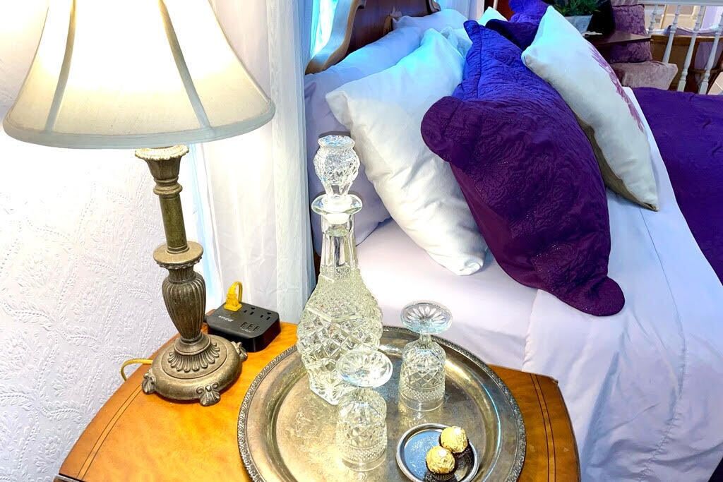 A bed with a purple blanket, a bedside table with a lamp, a tray with crystal glasses and chocolates, and a decorative bottle.