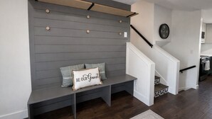Un intérieur moderne avec un banc gris, un coussin orné de l’inscription « SOYEZ LES BIENVENUS », une horloge et un escalier menant à la cuisine.