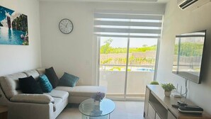 Ein modernes Wohnzimmer mit einer Couch, einem Glastisch, einem Fernseher und einem großen Fenster mit Blick auf Grünflächen.