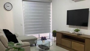 Ein modernes Wohnzimmer mit einer Couch, einem Glastisch, einem Fernseher und einem Fenster mit Jalousien.