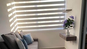 Ein modernes Wohnzimmer mit einem grauen Sofa, einem kleinen Beistelltisch mit Topfpflanze und einem Fenster mit horizontalen Rollläden.