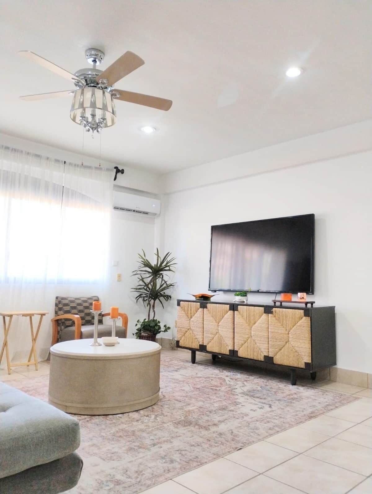 A modern living room with a ceiling fan, a flat-screen TV, a round ottoman, a sofa, a small table, and a potted plant.