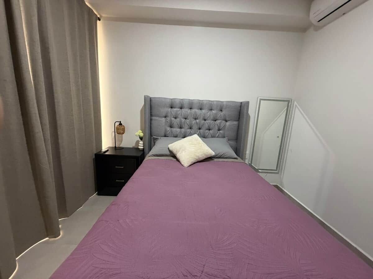 A bedroom with a tufted headboard, a bed with a purple bedspread, a bedside table with a lamp, and a nightstand.
