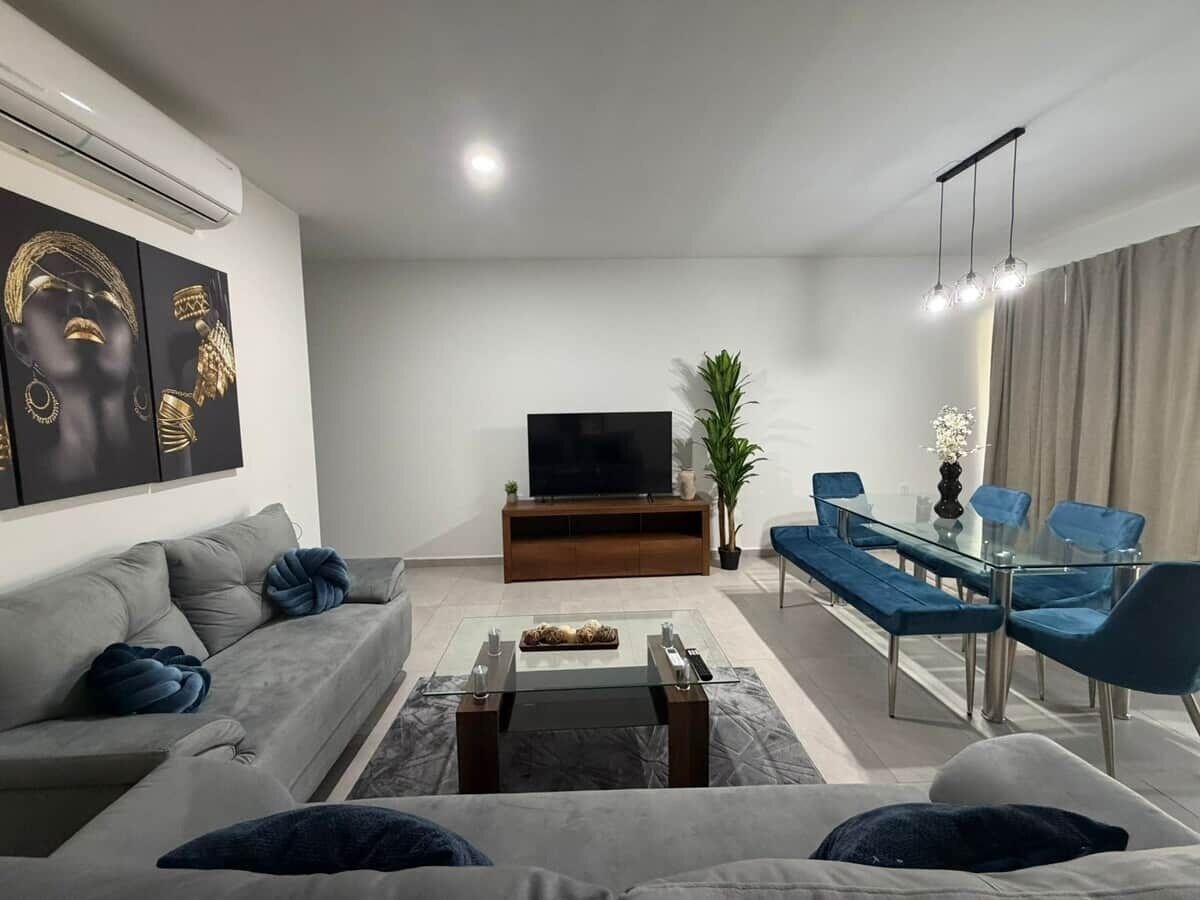A modern living room with a grey sofa, a glass coffee table, a TV on a wooden stand, and a dining area with blue chairs and a glass table.