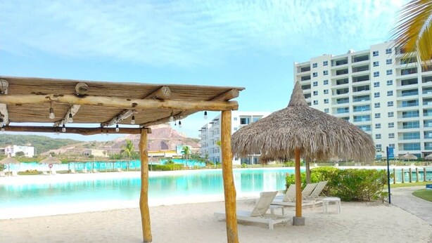 Ein Poolbereich am Strand mit einem Reetdachschirm, Liegestühlen und einer Holzpergola.