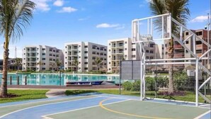 A basketball court with a net, palm trees, a swimming pool, and multi-story buildings in the background. - Lagoon view apartment plus exclusive amenities (Mazatlán)