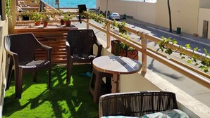 Een balkon met rieten meubels, een kleine tafel en potplanten. Achtergrond is een uitzicht op de oceaan en palmbomen.