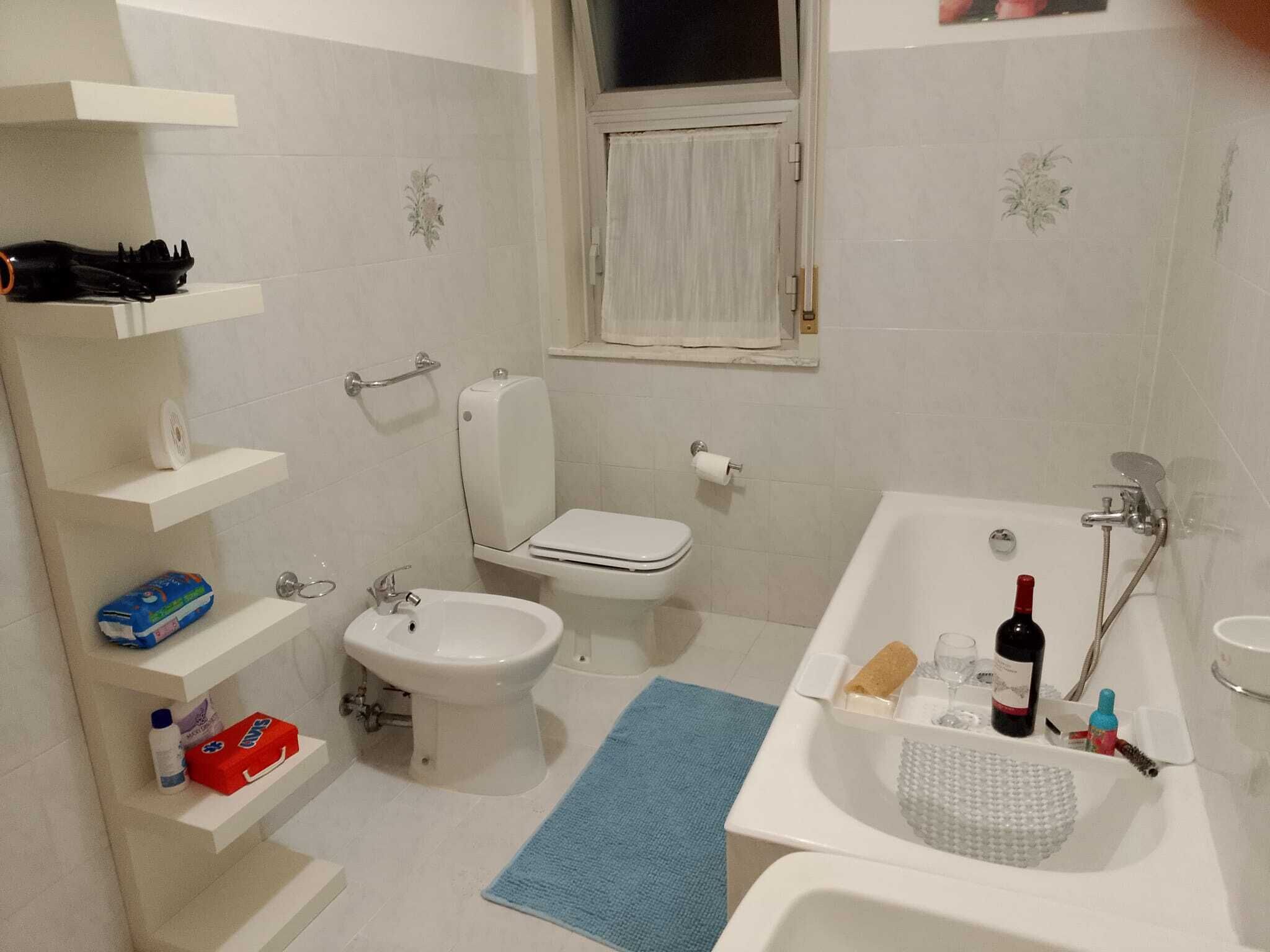 A bathroom with a toilet, bidet, sink, and bathtub. There are shelves with toiletries and a window above the toilet.