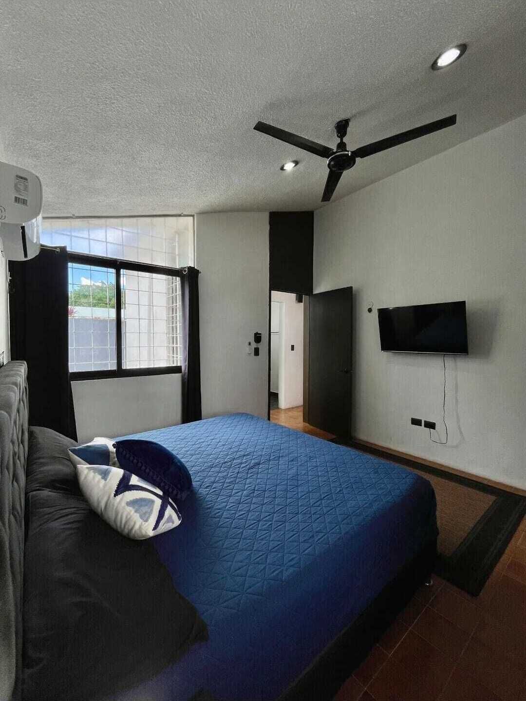A bedroom with a bed, a ceiling fan, a TV mounted on the wall, and a window with curtains.