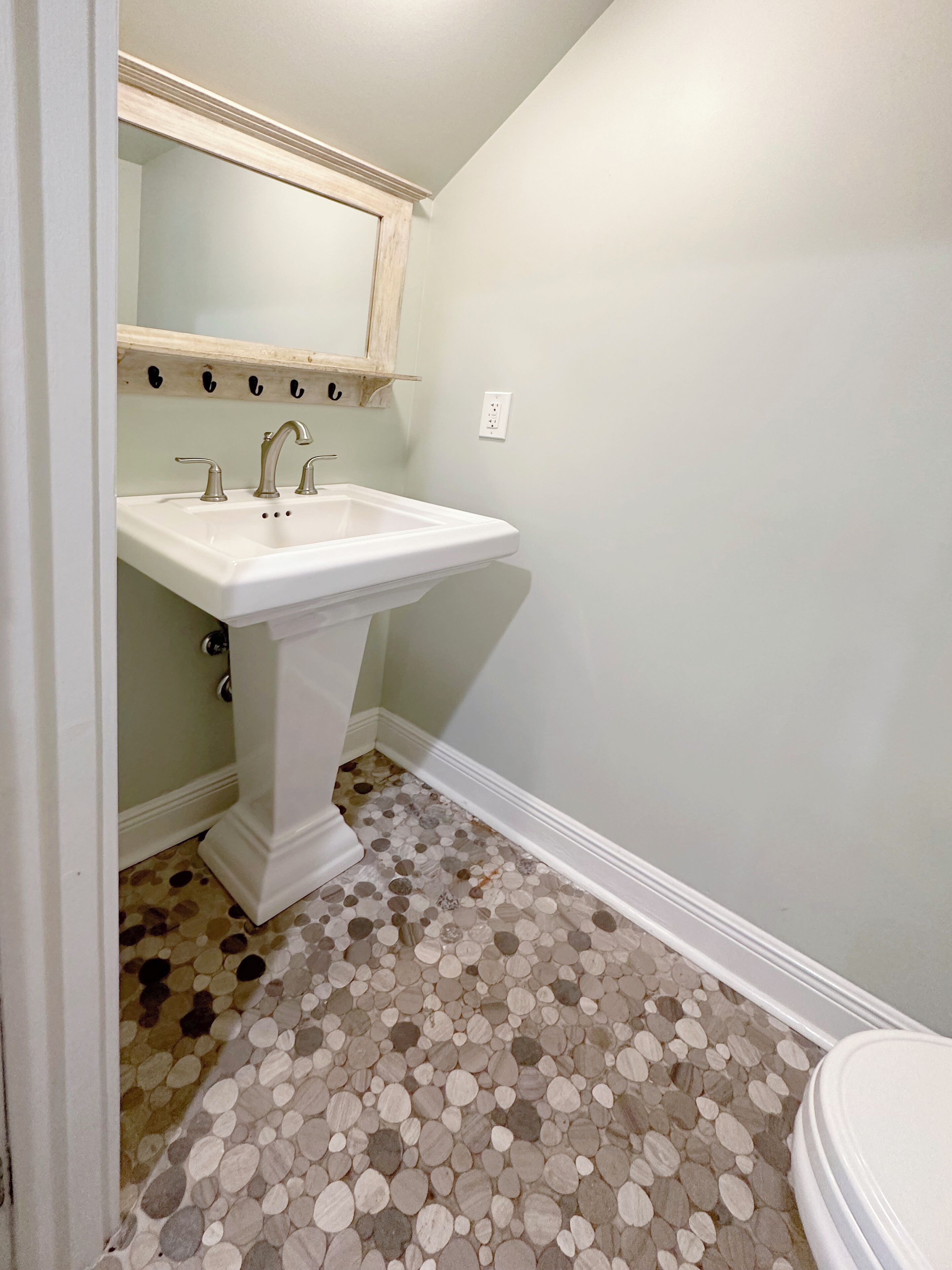 Une salle de bain avec un lavabo sur colonne, un miroir au-dessus et des toilettes. Le sol est recouvert d’un motif imitant des galets.