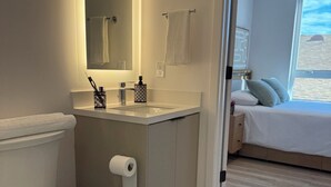 Ein Badezimmer mit Toilette, Waschbecken und Spiegel. Im angrenzenden Schlafzimmer befindet sich ein Fenster.