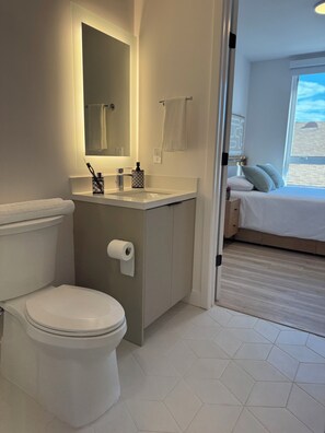 Ein Badezimmer mit Toilette, Waschbecken und Spiegel. Im angrenzenden Schlafzimmer befindet sich ein Fenster.