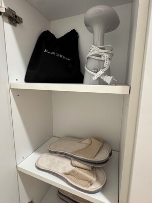 Ein Lagerraum mit einer schwarzen Tasche mit dem Aufdruck „Haartrockner“ und einem weißen Haartrockner auf der obersten Ablage, sowie beigen Hausschuhen auf der untersten Ablage.