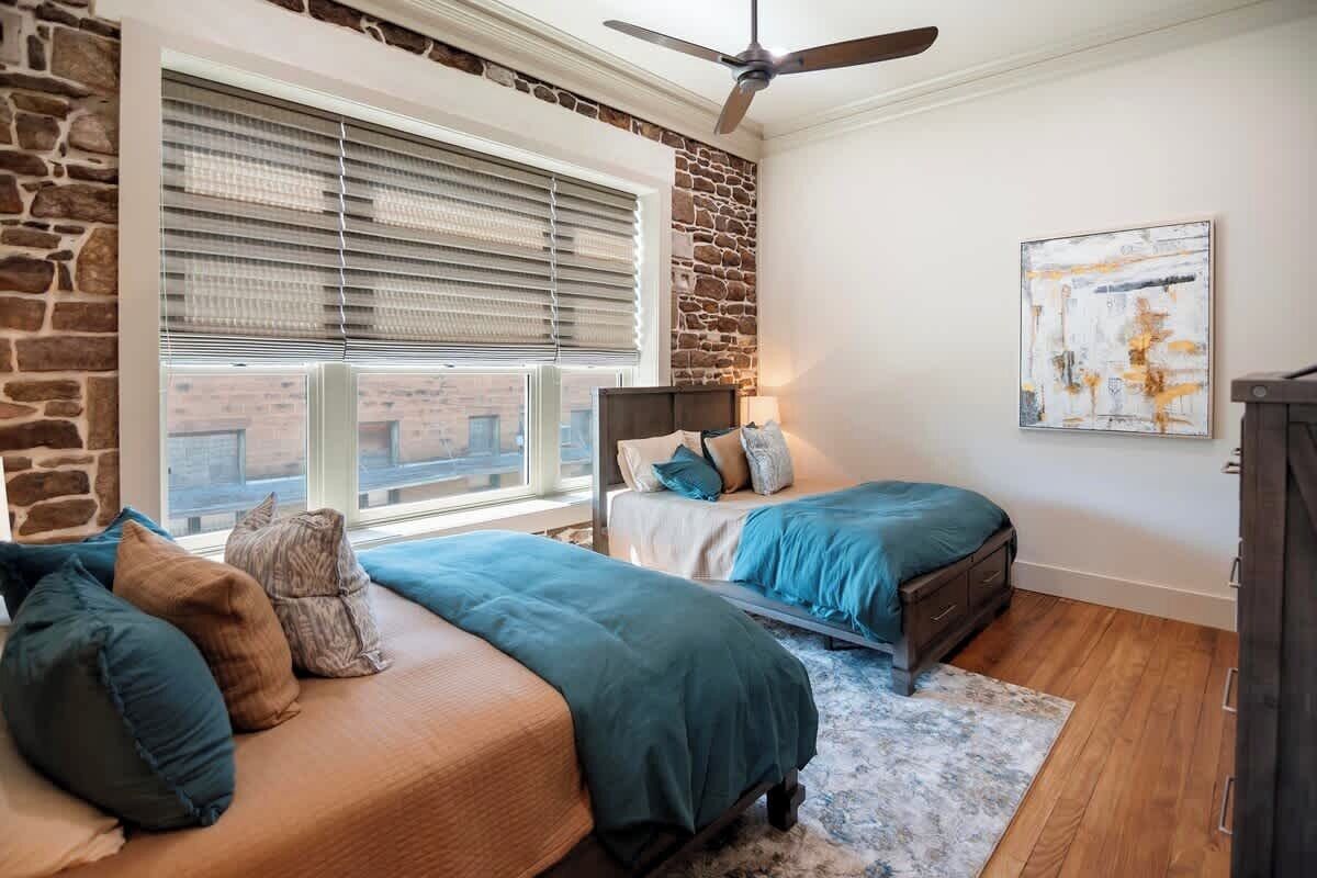 A bedroom with two beds, a stone accent wall, a ceiling fan, and a large window with blinds.