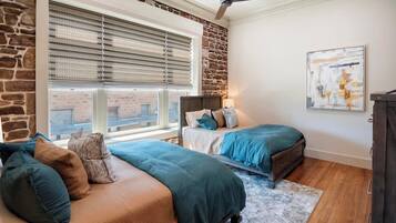 A bedroom with two beds, a stone accent wall, a ceiling fan, and a large window with blinds.