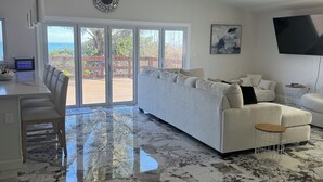 A spacious living area with a large white sofa, a dining table with chairs, and a kitchen area with a TV and a decorative wall piece. - Stunning Ocean Views! 2-bedroom house, private beach in Melbourne Beach, FL (Melbourne Beach)
