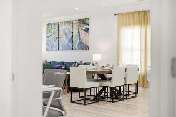 A modern dining area with a round table, white chairs, and a green sofa in the background. - Waves on Walnut NEW 3BR Home w/Private Pool in CG (North Myrtle Beach)
