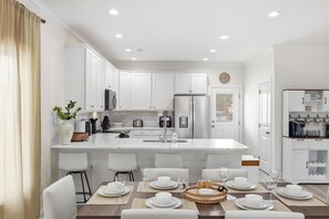 A modern kitchen with white cabinetry, a central island, and a dining area set for four. - Waves on Walnut NEW 3BR Home w/Private Pool in CG (North Myrtle Beach)