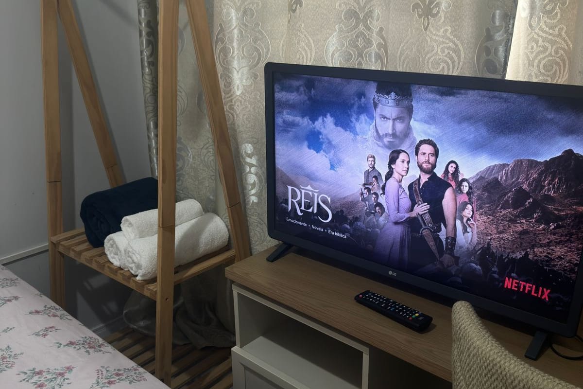 Quarto de hotel com uma cama, uma estante de madeira com uma televisão, um controle remoto e um cabideiro.