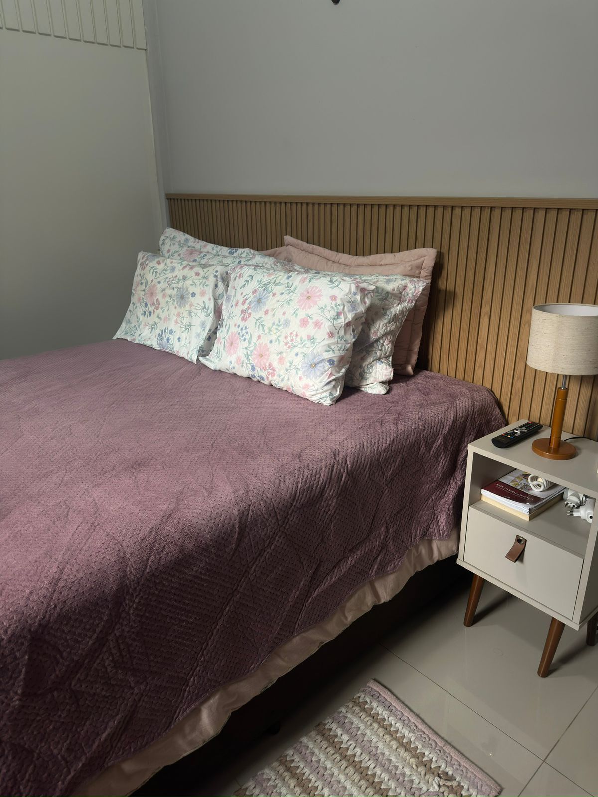Ein ordentlich bezogenes Bett mit violettem Überwurf und blumigen Kissen, ein Nachttisch mit Lampe und Fernbedienung sowie ein Teppich auf dem Boden.