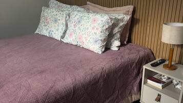 Ein ordentlich bezogenes Bett mit violettem Überwurf und blumigen Kissen, ein Nachttisch mit Lampe und Fernbedienung sowie ein Teppich auf dem Boden.