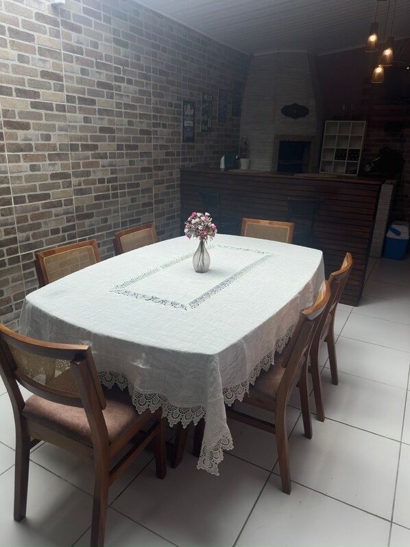 Una mesa de comedor redonda con mantel de encaje blanco, rodeada de sillas de madera con asientos acolchados. Un jarrón con flores está colocado en el centro.