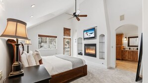 A bedroom with a bed, a fireplace, a TV, and a kitchen area visible through an open door. - Spruce Street Chalet | Cozy Mountain Living (Vail)