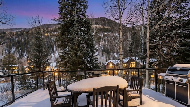 A snow-covered deck with a table and chairs, overlooking a mountainous area with houses in the background. - Spruce Street Chalet | Cozy Mountain Living (Vail)