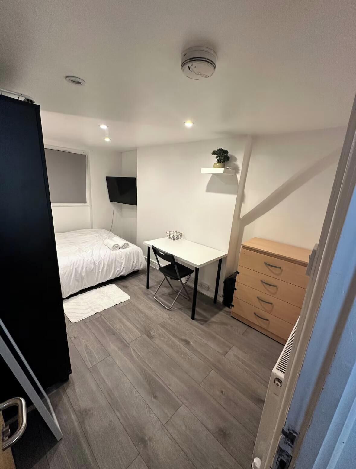 Ein kleines Schlafzimmer mit Bett, Schreibtisch, Kommode und einem an der Wand befestigten Fernseher.