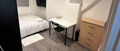 Ein kleines Schlafzimmer mit Bett, Schreibtisch, Kommode und einem an der Wand befestigten Fernseher.