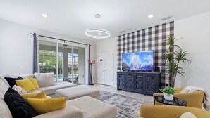 A modern living room with a sofa, ottoman, and armchair, a flat-screen TV, a dark wooden cabinet, and a large window with a view of greenery. - Enchanted Palms Villa 7BR w Private Pool (Davenport)