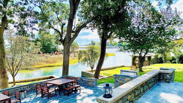 An outdoor patio with stone flooring, wooden furniture, and a view of a lake. - Luxury Waterfront Estate · Sleeps 18 · 45-Min Drive to FIFA World Cup Stadium (East Rutherford)