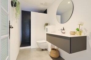 Casa de banho moderna com sanita branca, toucador escuro, espelho redondo e planta pendente.
