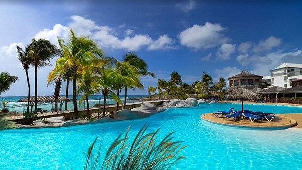 Una piscina de resort con palmeras, una playa y un edificio de fachada blanca.