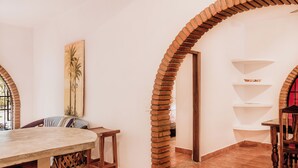 A dining area with a long table, wooden chairs, and a brick column. There are shelves with decorative items and a painting of palm trees. - Casa Roja · Whole Home with Pool, Terrace & Palapa · Local Sayulita (Sayulita)