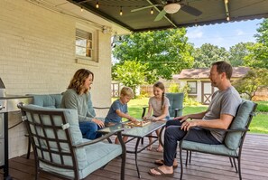 Une famille de quatre personnes est assise à une table à l’extérieur, sur une terrasse couverte, avec un mur en briques et une plateforme en bois.