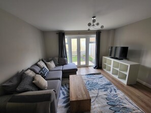 A living room with a sectional sofa, a wooden coffee table, a TV on a white cabinet, and a large window with curtains.