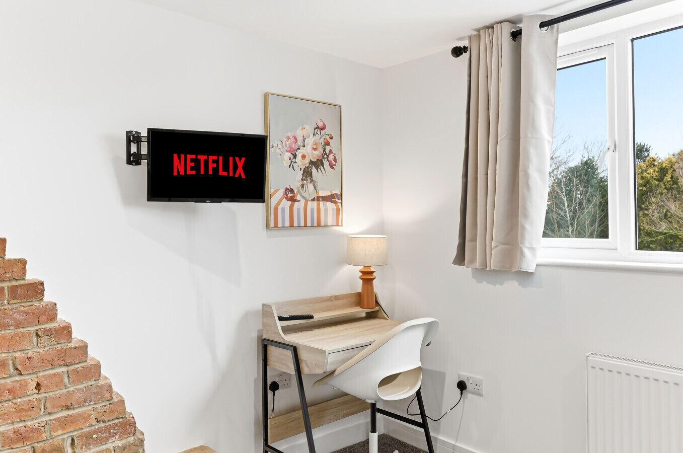 A room with a desk, a chair, a lamp, a TV mounted on the wall, and a window with curtains.