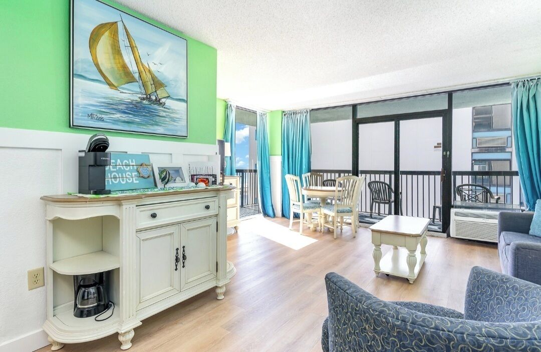 A living room with a dining area, a large window with curtains, a painting of a sailboat, and a kitchenette with a coffee maker.