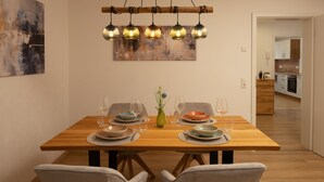 Un comedor con una mesa de madera preparada para dos personas, rodeada de sillas de mimbre. Una lámpara colgante moderna ilumina la mesa, y se aprecia un cuadro en la pared a la izquierda.