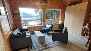 A cozy wooden cabin interior with a sofa, armchairs, a coffee table, and a large window offering a view of a lake and mountains. - Remote Alaska cabin, located in the Kachemak Bay Park area of Tutka Bay. (Seldovia)