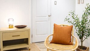 Una habitación con una puerta blanca, una silla de mimbre con cojín marrón, una mesita con una lámpara y un cuenco, y una planta en maceta.