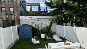 Un patio trasero con una mesa y sillas blancas, un cobertizo azul y una cerca blanca.
