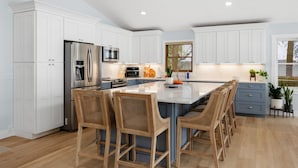 A modern kitchen with white cabinets, stainless steel appliances, a central island with bar stools, and a view of the outdoors through a window. - Long Lake Haven | 4BR | Waterfront | Boat Rental (Traverse City)
