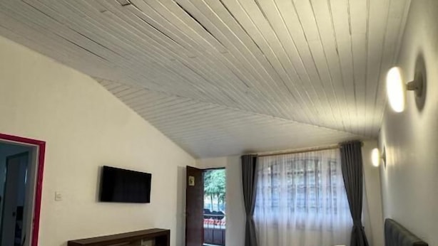 Un dormitorio con cama, televisor montado en la pared, techo de madera y ventana con cortinas.