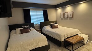 Ein Hotelzimmer mit zwei Betten, einer Bank, einem Fenster mit Jalousien und gerahmten Bildern an der Wand.